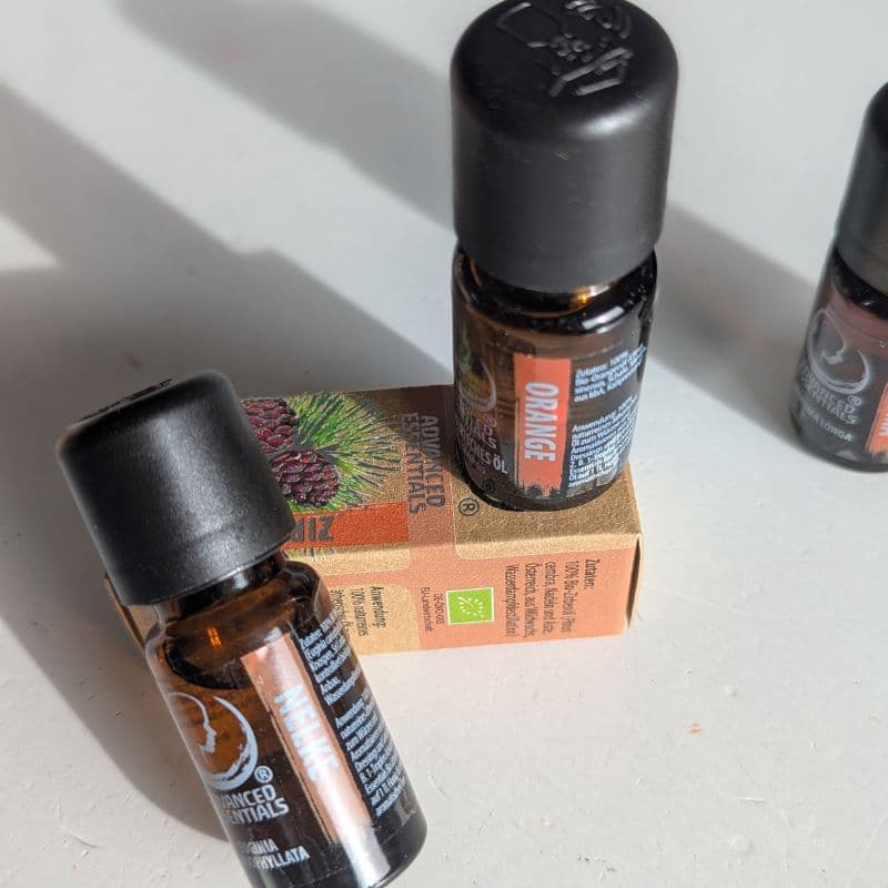 Zwei Fläschchen ätherischer Öle von Advanced Essentials – Orange und Nelke – auf einer Verpackung mit Kiefernzapfen-Motiv, fotografiert im warmen Tageslicht.
