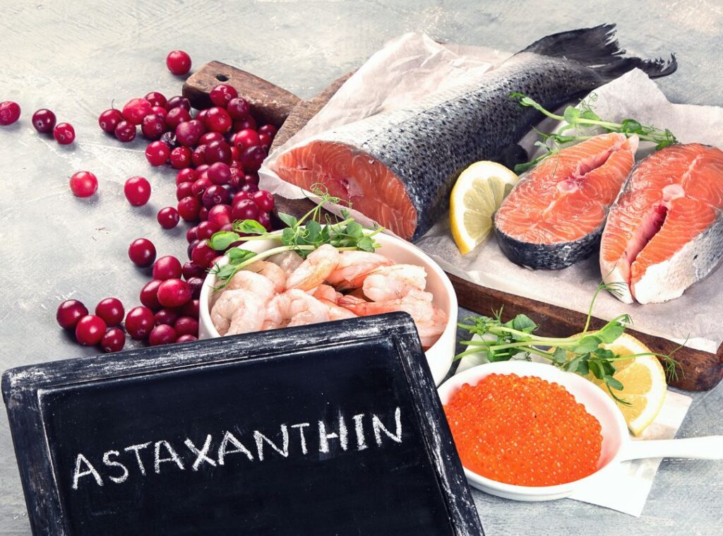 Natürliche Astaxanthin-Quellen aus der Ernährung: Lachs, Garnelen, Beeren und Meerestierprodukte. Auf dem Bild liegen frische Lebensmittel, die natürlicherweise Astaxanthin enthalten: Lachsfilets, Garnelen, roter Fischrogen und leuchtende Beeren. Im Vordergrund steht eine kleine Tafel mit der Aufschrift „Astaxanthin“.