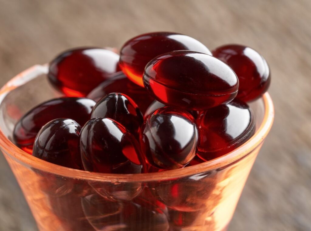 Astaxanthin in Kapselform: Die charakteristische rote Farbe stammt aus Mikroalgen. Nahaufnahme von roten Astaxanthin-Kapseln in einem kleinen Glasbehälter. Die glänzenden Softgel-Kapseln zeigen die typische dunkelrote Farbe des natürlichen Carotinoids.