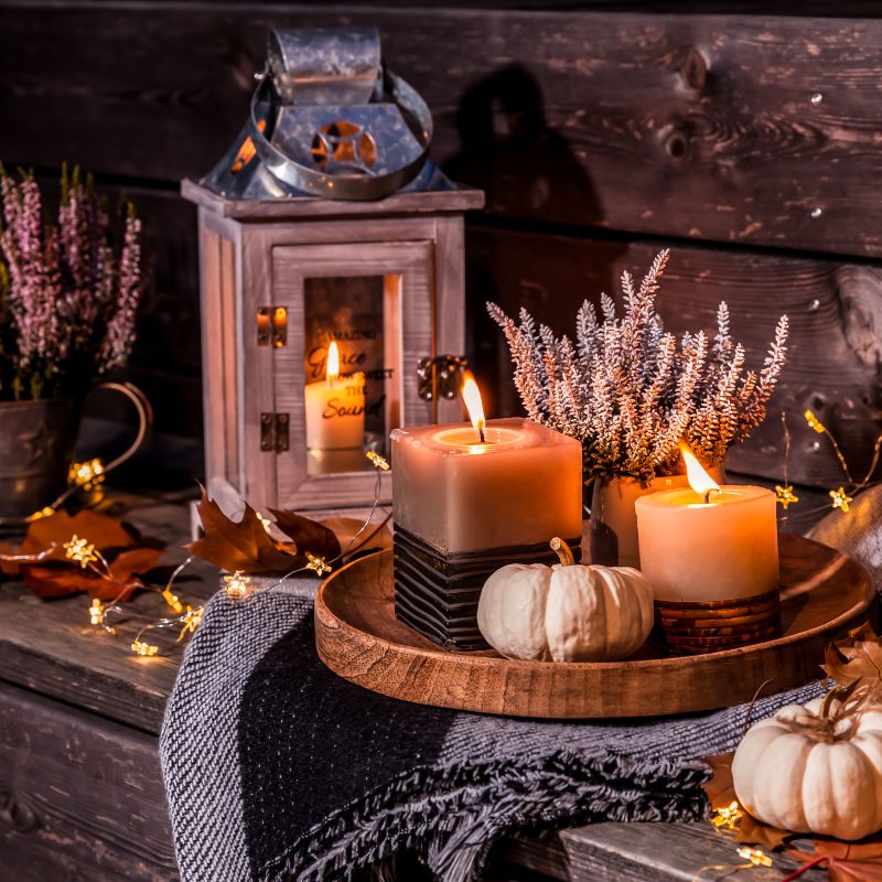 Gemütliche Herbstdeko mit brennenden Kerzen, kleiner Laterne, weißen Zierkürbissen, Heidekraut und Lichterkette auf rustikalem Holztisch