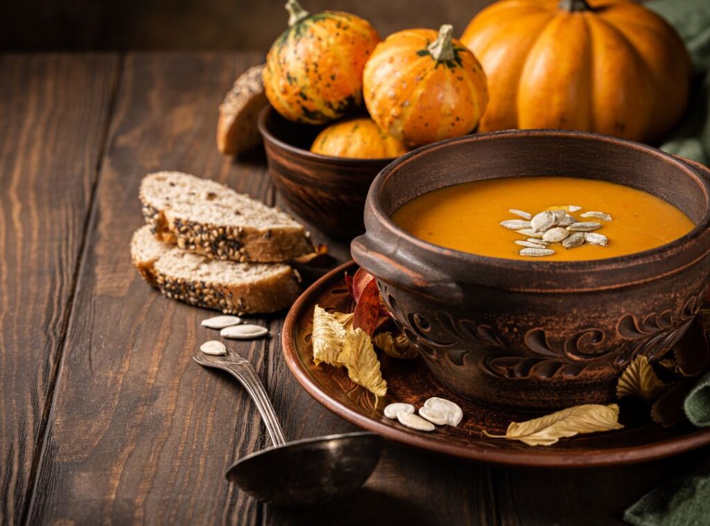 Herbstliche Kürbiscremesuppe in rustikaler Schale, dekoriert mit Kürbiskernen, serviert mit frischem Körnerbrot und Mini-Kürbissen auf Holztisch.